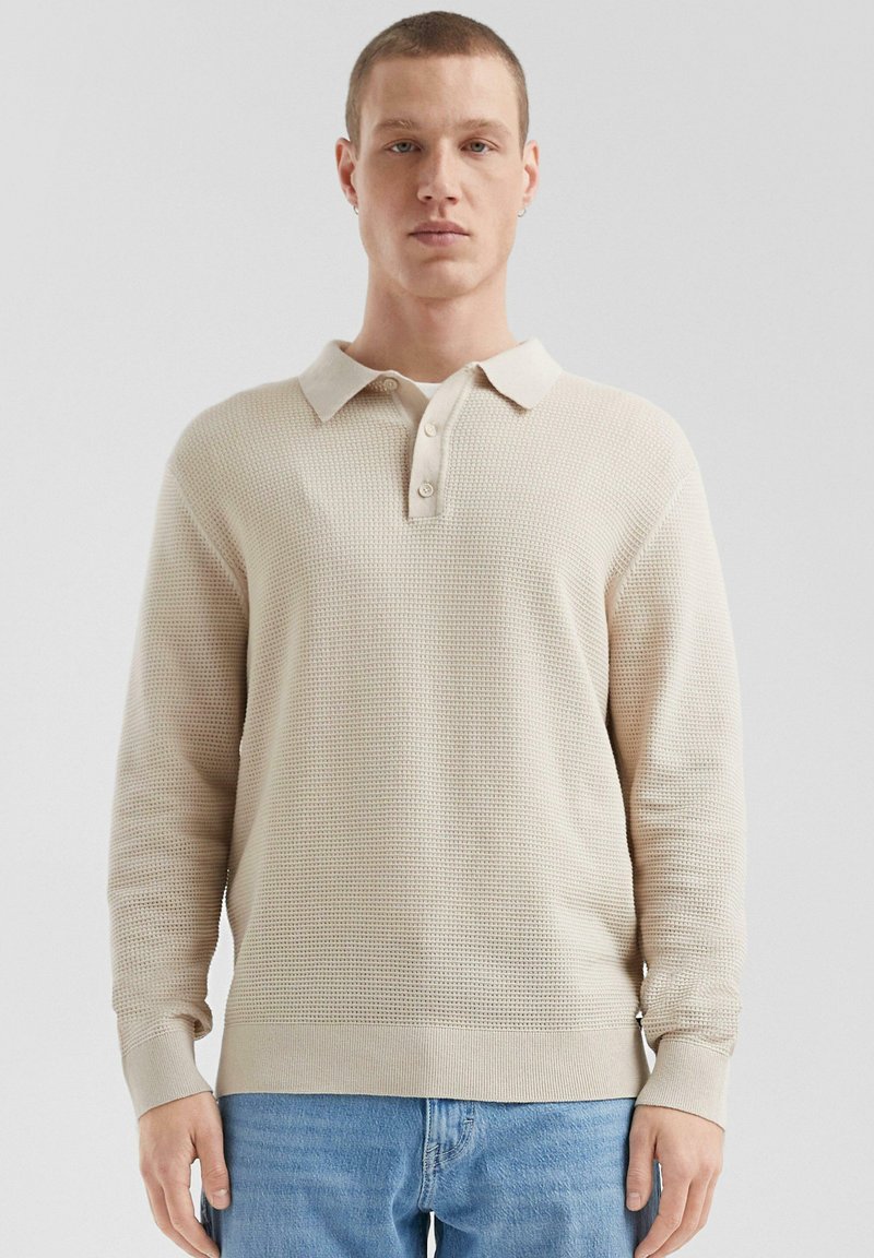 Beige strukturierte langärmlige Poloshirt mit kragenförmigem Ausschnitt und drei Knöpfen, mit gerippten Bündchen und Saum. Kombiniert mit hellblauen Jeans.