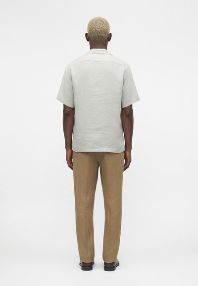 Άνδρας με κοντά ξανθά μαλλιά, που φοράει ανοιχτό γκρι κοντομάνικο πουκάμισο, καφέ παντελόνι και μαύρα παπούτσια, στέκεται με την πλάτη στον θεατή.