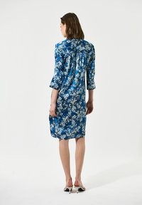 Abito in satin blu con motivo floreale, maniche a tre quarti, dettaglio arricciato sulla schiena e taglio al ginocchio. Indossato con sandali open-toe.