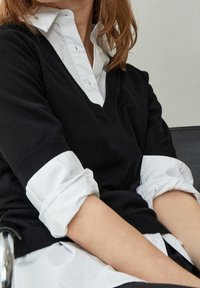 Personne assise, les bras croisés, portant un pull noir à encolure en V par-dessus une chemise blanche boutonnée aux manches retroussées.