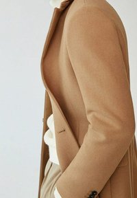 Lysebrunt uldblandingsfrakke med klippede slag, enkelt knapdetalje og skræddersyet pasform. Tekstureret stof og sideåbning fremhæver designet.