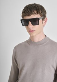 Giovane uomo dai capelli ricci corti, che indossa occhiali da sole rettangolari neri oversize e un maglione beige a collo alto, rivolto in avanti.