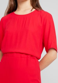Vestido rojo de manga corta con escote redondo y cintura fruncida. La tela parece lisa y ligera, con un ajuste holgado.