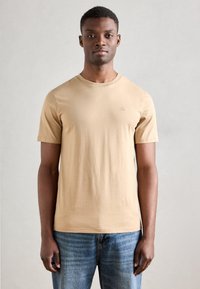 Camiseta de algodón beige con cuello redondo y mangas cortas; presenta un pequeño logo en la parte izquierda del pecho; combinada con jeans azules.