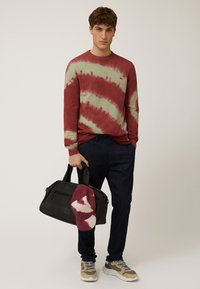 Rode en groene tie-dye sweatshirt, marineblauwe broek, een zwarte sporttas met een rood-witte pet erop. Schoenen zijn beige en grijs.