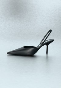 Μαύρο γυναικείο παπούτσι με λεπτό στιλέτο τακούνι, μυτερή μύτη και διχτυωτό λουράκι, σε ανοιχτό γκρι φόντο και επιφάνεια.