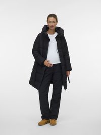 Grūtniece, ģērbusies melnā pildītā mētelī, baltā krekā, melnās biksēs un brūnās zābakos, stāv pretī vienkāršam baltam fona.