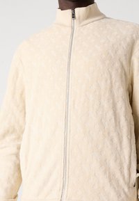 Ljust beige jacka med full dragkedja och subtilt texturerat geometriskt mönster, ribbad krage och dragkedjefickor, bärs av en person.