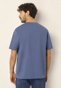 Modré tričko s krátkým rukávem, kulatým výstřihem a hladkým povrchem. Zdá se, že je obepnuté na ramenou, volné kolem pasu.