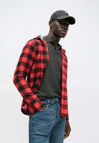 Camisa de flanela xadrez vermelho e preto com um polo cinza por baixo, combinada com jeans azuis e um boné de basebol de denim escuro.