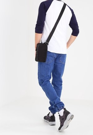 Zwarte stoffen crossbody tas met een logo, met een rechthoekige vorm en een verstelbare schouderband, gedragen over een blauwe shirt en jeans.