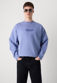 Lila pamut kapucnis pulóver, kerek nyakkal, sötétkék "BARBOUR INTERNATIONAL" felirattal. Fekete farmernadrággal viselve.