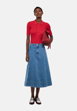 Femme portant un polo rouge à manches courtes, une longue jupe en jean bleue, des chaussures blanches et bordeaux, tenant un sac à main rouge tissé.