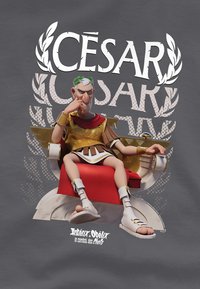 Karikaturhafte Darstellung von Caesar in einer goldenen Toga und rotem Umhang, sitzend auf einem Thron mit einem roten Kissen, vor einem grauen Hintergrund.