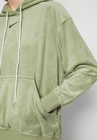 Zaļš velūra hoody ar auklītēm, priekšējo kabatu, ribotām aprocēm un smalku Nike logo tumšā krāsā. Mīksta tekstūra un brīvs piegriezums.