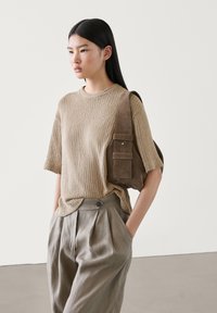 Beige ribbed knit sweater with short sleeves, paired with light gray pleated trousers. The model carries a brown suede shoulder bag with pockets.
