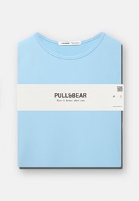 T-shirt azzurro chiaro realizzato in cotone, con scollo rotondo e maniche corte, piegato con una fascia di carta bianca che mostra il marchio.