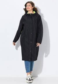 Svart vattentät lång jacka med huva och neongrön foder, med tryckknappar, sidofickor och en avslappnad passform. Bärs över jeans.