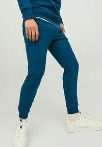 Pantaloni jogger teal in morbido tessuto con polsini elasticizzati, caratterizzati da una vestibilità slim e tasche laterali, abbinati a sneakers bianche.