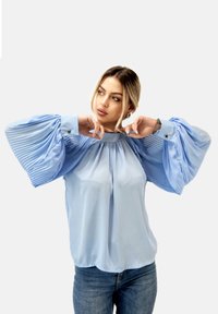 Lichtblauwe blouse met geplooide, volumineuze mouwen en een ronde halslijn. Heeft knoopsluiting bij de manchet en een zachte, vloeiende stof.