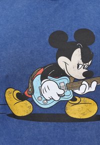 Blå t-shirt med en stor grafik av en tecknad mus som spelar en ljusblå gitarr, med gula skor och svarta detaljer på figuren.