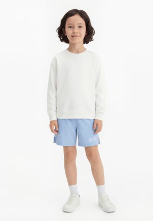 Dieťa s tmavými vlasmi po plecia, oblečené v bielom tričku s dlhým rukávom, svetlomodrých šortkách, bielych ponožkách a bielych teniskách, stojace nehybne.