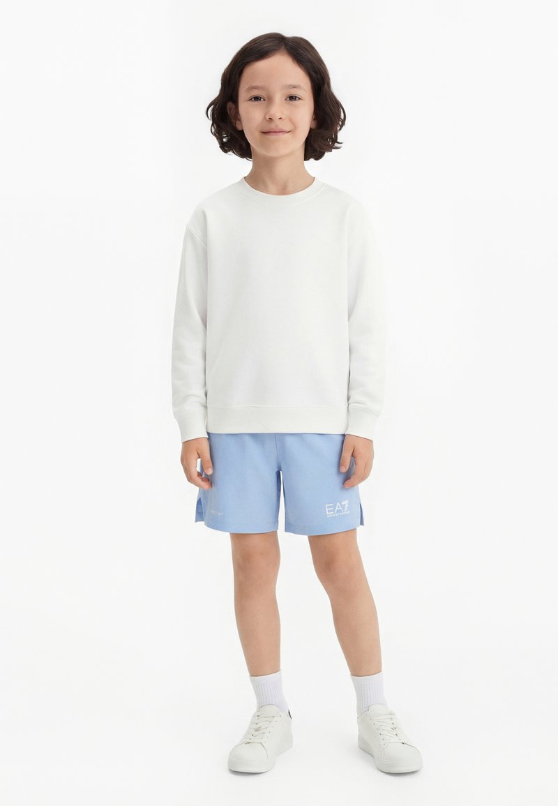 Παιδί με σκούρα μαλλιά μέχρι τους ώμους, που φορά λευκό μπλουζάκι με μακριά μανίκια, ανοιχτό μπλε σορτς, λευκές κάλτσες και λευκά αθλητικά παπούτσια, στέκεται ακίνητο.