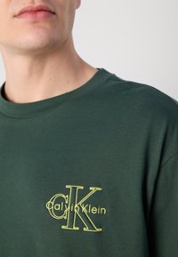 Vihreä puuvillapaita, jossa on pyöreä kaula-aukko. Paidassa on keltaisella brodeerattu "Calvin Klein" -logo tyylitellyssä fontissa vasemmassa rintataskussa.