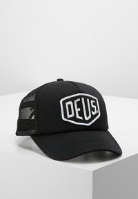 Gorra negra con visera redondeada, que cuenta con una parte frontal de tela sólida y paneles laterales de malla. En la parte frontal se muestra una insignia de logo bordado en blanco.