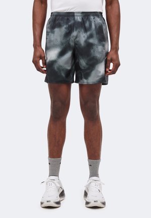 Mann trägt schwarze und graue sportliche Shorts mit Muster, graue Kniestrümpfe und weiße Nike-Sneaker und steht vor einem schlichten Hintergrund.