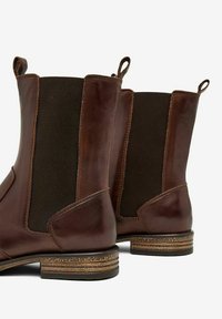 Bottines en cuir marron avec panneaux élastiques sur les côtés, finition texturée et languettes de tirage. Talon empilé notable et accents noirs contrastants.