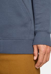 Højre hånd hviler ved siden, iført en blå sweatshirt med ribbede manchetter og talje over sennepsfarvede bukser.