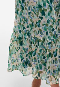 Orlo dell'abito con dettaglio di volant in motivo astratto a pennellate nei toni del verde, blu e beige su tessuto leggero e semitrasparente.