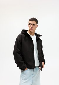 Schwarze Zip-Jacke mit Kapuze, mit einer glatten Textur und seitlichen Taschen, getragen über einem weißen T-Shirt und hellblauen Jeans.