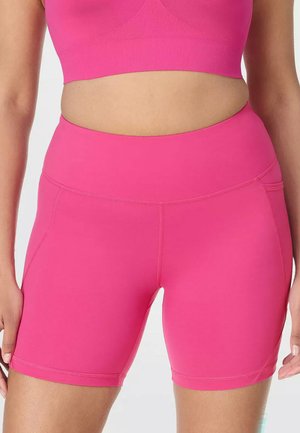 Vrouw draagt felroze atletische shorts met hoge taille en bijpassende sportbeha, getoond van de buik tot halverwege de dij, handen ontspannen langs het lichaam.