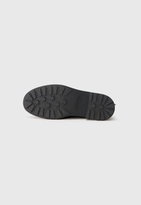 Suela de zapato negra con surcos profundos y empuñaduras en forma de estrella, plana contra un fondo claro liso, mostrando el nombre de la marca grabado.