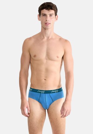 Młody mężczyzna z krótkimi kręconymi włosami, ubrany w niebieskie majtki Sloggi, stojący na tle jednolitego białego tła, zwrócony przodem.