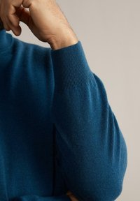 Bras droit plié, main près du visage, portant un pull bleu à manches longues avec des poignets côtelés, sur un fond neutre.