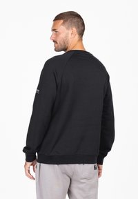 Sudadera negra con mangas largas, cuello redondo, puños y dobladillo de canalé. Presenta una pequeña etiqueta en la manga con detalles de texto.