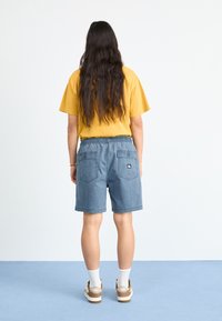 Gul t-skjorte og blå shorts med elastisk midje. Shortsene har lommer og et logo-patch. Modellen har på seg hvite sokker og lysebrune sko.