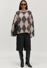 Vrouw in een oversized roze en grijze ruittrui, knielange zwarte shorts, zwarte hoge hakken, zonnebril en met een zwarte clutch in haar hand.
