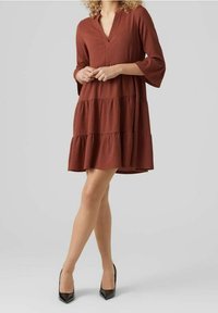 Vestido en tono óxido, de capas, con escote en V y mangas tres cuartos, hecho de una tela texturizada. Combinado con zapatos de tacón negro.