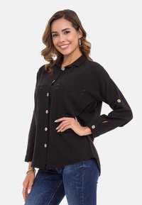 Zwarte blouse met knopen van stof met een kraag, voorzien van twee borstzakken, houten knopen en details op de manchetten. Draagt met blauwe spijkerbroek.