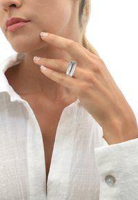 Anello d'argento con design intrecciato indossato su un dito, con uno sfondo di una camicia bianca con colletto e tono della pelle. Texture semplici e lisce.