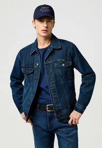 Donkerblauwe spijkerjas met knoopsluiting aan de voorkant, twee borstzakken en metalen hardware. Draagt over een marineblauwe t-shirt en bijpassende jeans. Geaccentueerd met een donkere pet.