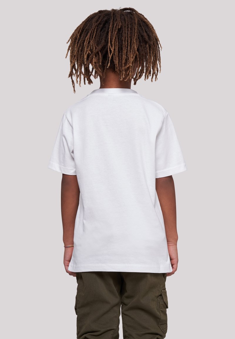 Enfant avec des dreadlocks brunes portant un t-shirt blanc uni à manches courtes et un pantalon cargo vert foncé, dos tourné sur un fond uni.
