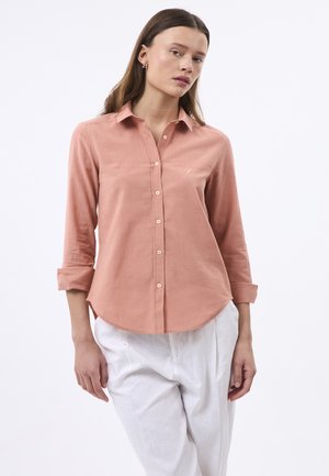 Hedvábná button-up košile v broskvové barvě s dlouhými rukávy, vyrobená z měkké tkaniny. Zaoblený lem a malý logotyp Detail na hrudi. V kombinaci s bílými kalhotami.