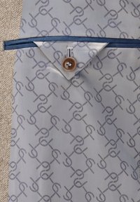 Blau gemusterter Stoff mit einem wiederholten Logodesign, versehen mit einem braunen Knopf und einem kleinen Reißverschlussdetail, neben einem strukturierten beigen Material.