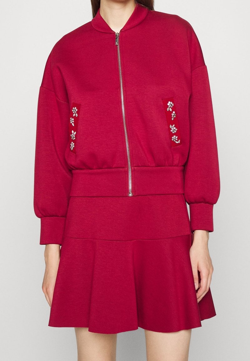 Czerwona bomberka z zapięciem na zamek i ozdobnymi kwiatami na kieszeniach. Rozkloszowana spódnica o gładkiej fakturze, podkreślająca wąską talię i luźny dół.