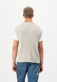 Gilet en tricot rayé beige avec un col rond, superposé à un T-shirt blanc, présentant une texture côtelée et une étiquette avec logo à l'arrière.